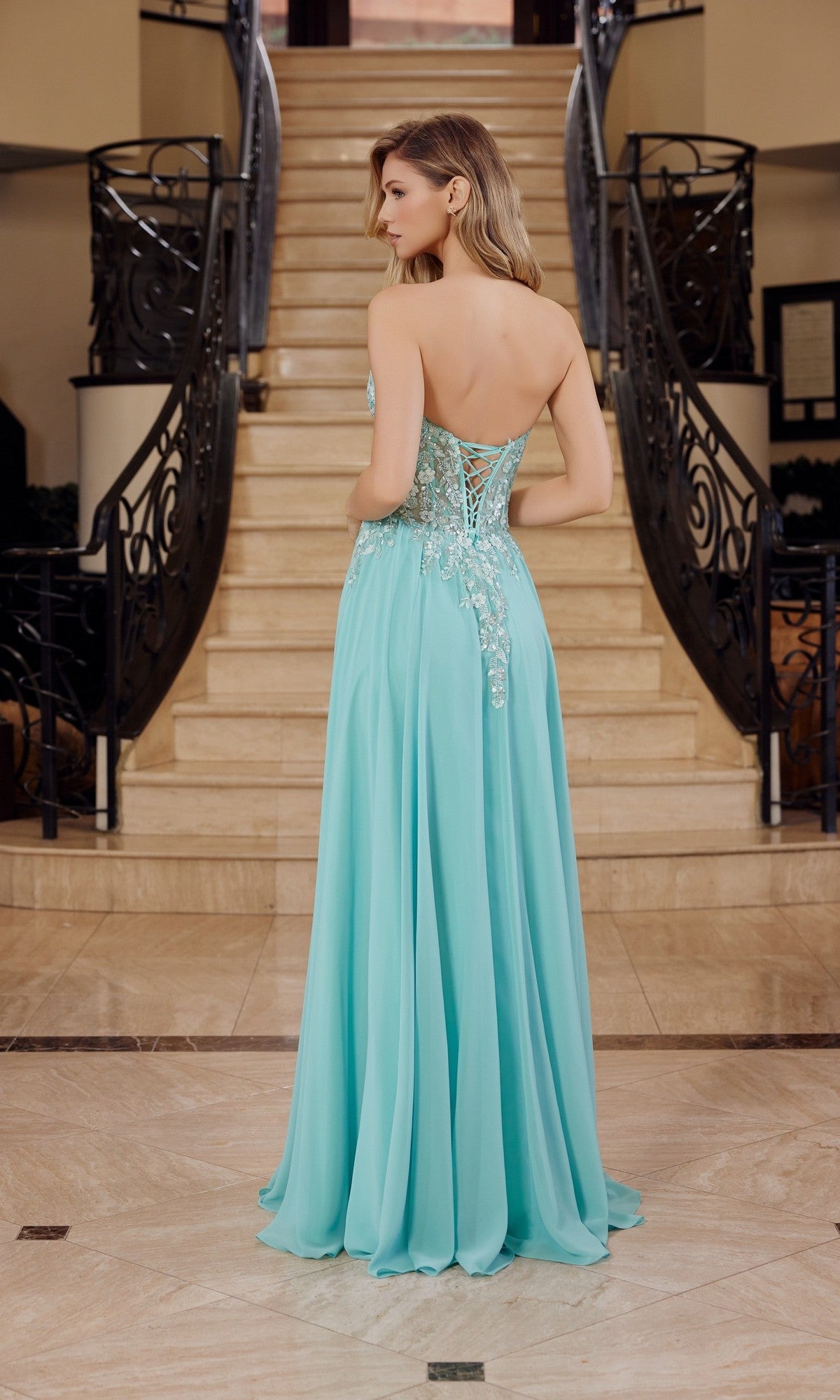 A woman in the Nox Anabel C1617 Strapless Sweetheart Prom Dress with delicate floral details stands at the base of a marble staircase in an elegant setting.